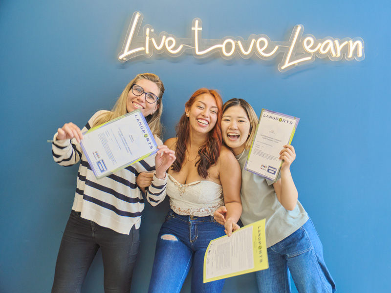 Drei Sprachschülerinnen halten stolz ihre Sprachzertifikate vor der Wand mit dem Motto „Live Love Learn“