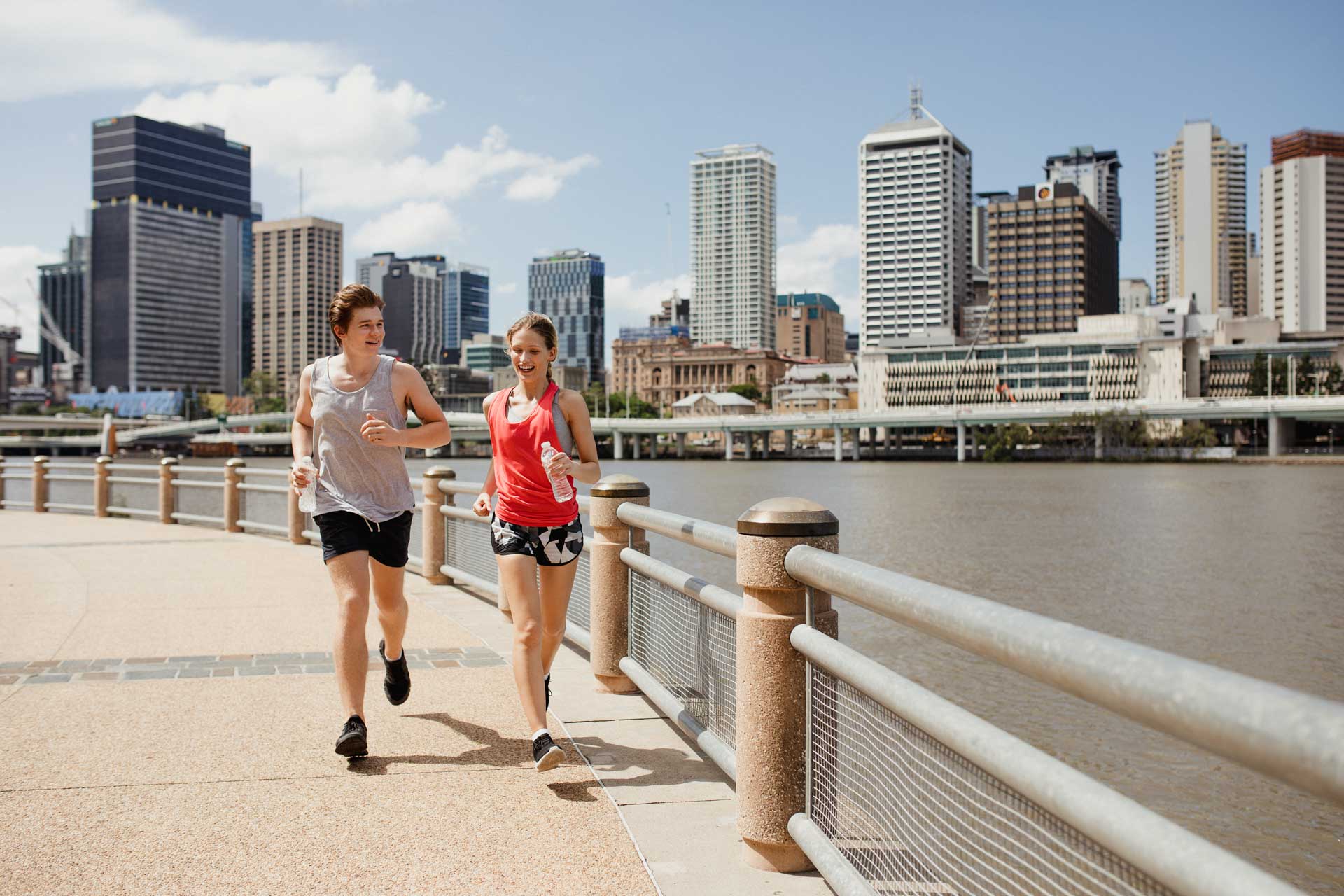Zwei junge Menschen joggen am Flussufer in Brisbane mit moderner Skyline im Hintergrund bei sonnigem Wetter.