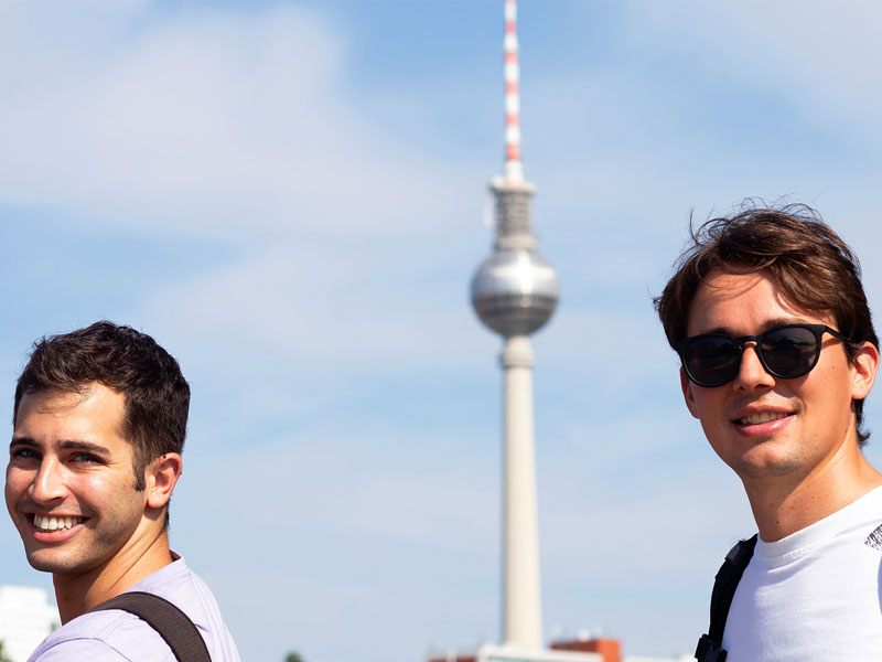 Zwei junge Erwachsene mit Rucksack vor dem Berliner Fernsehturm bei sonnigem Wetter