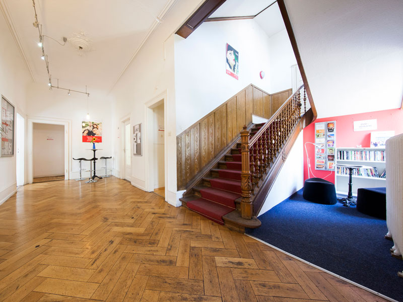 Heller Eingangsbereich mit Holztreppe und Informationsbereich in der Alpadia Sprachschule Freiburg