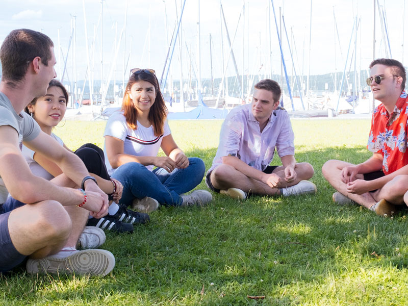 Gruppe junger Erwachsener sitzt auf einer Wiese am Hafen von Konstanz und unterhält sich in entspannter Atmosphäre