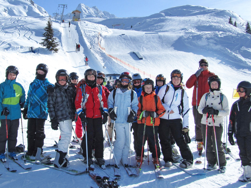 Gruppe junger Skifahrerinnen und Skifahrer posiert fröhlich im Schnee vor einer Skipiste in den Alpen