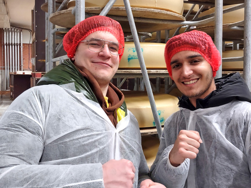 Zwei Teilnehmende in Schutzkleidung und roten Haarnetzen besuchen eine Käserei und lächeln in die Kamera