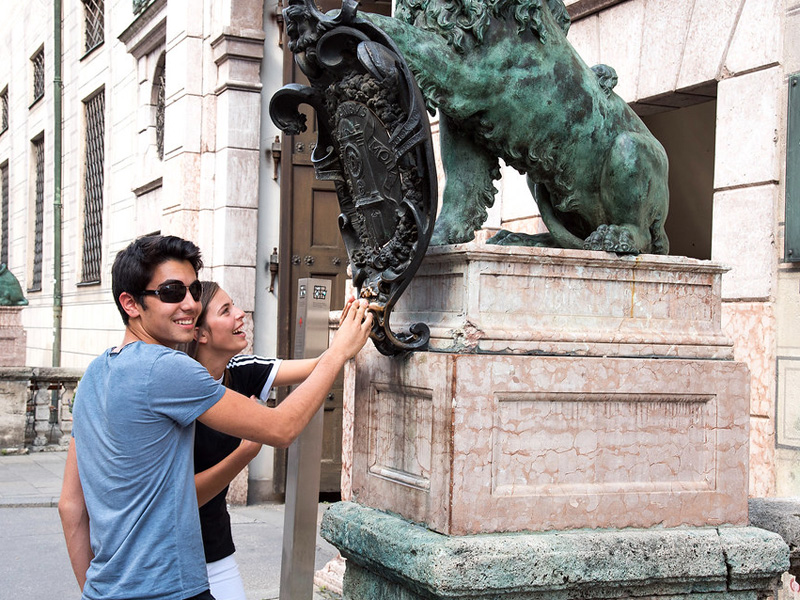 Zwei Jugendliche besichtigen eine Sehenswürdigkeit in München und berühren das Wappen einer Löwenstatue