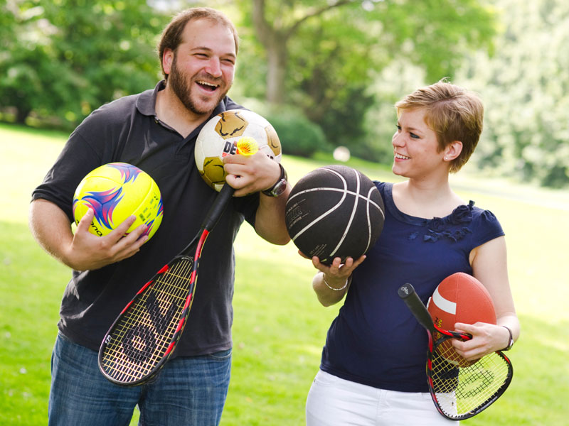 Zwei Personen mit verschiedenen Bällen und Schlägern bereiten sich lachend auf sportliche Aktivitäten im Park vor