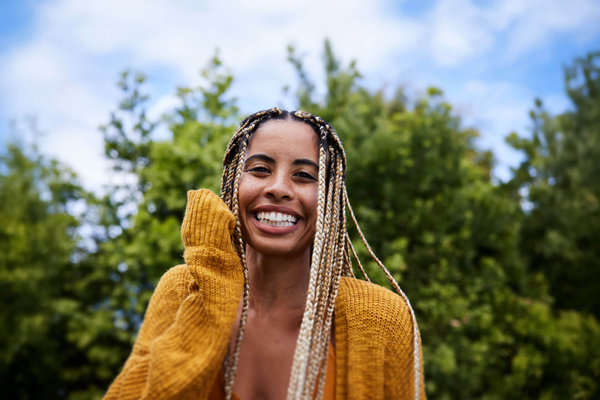 Lachende Person im gelben Pullover steht im Grünen unter blauem Himmel