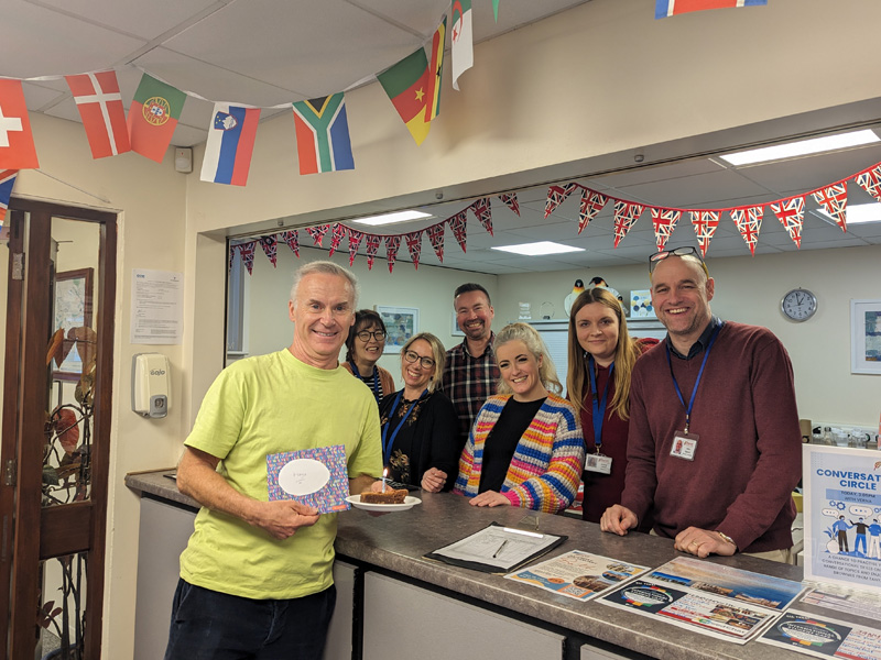Teammitglieder der Sprachschule posieren fröhlich hinter dem Empfangstresen, geschmückt mit Flaggen