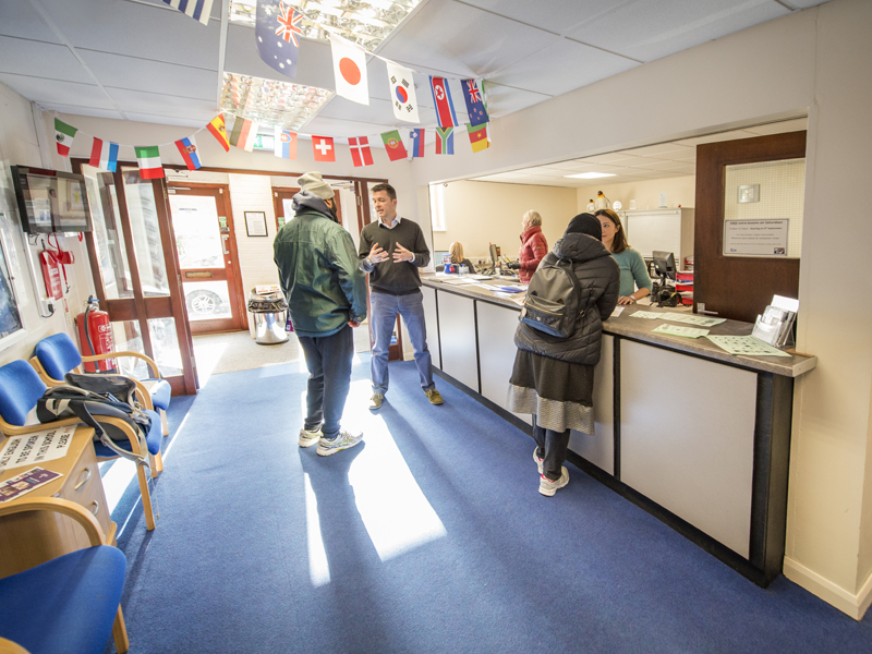 Empfangsbereich der Schule mit internationalen Flaggen und Studierenden im Gespräch mit dem Personal