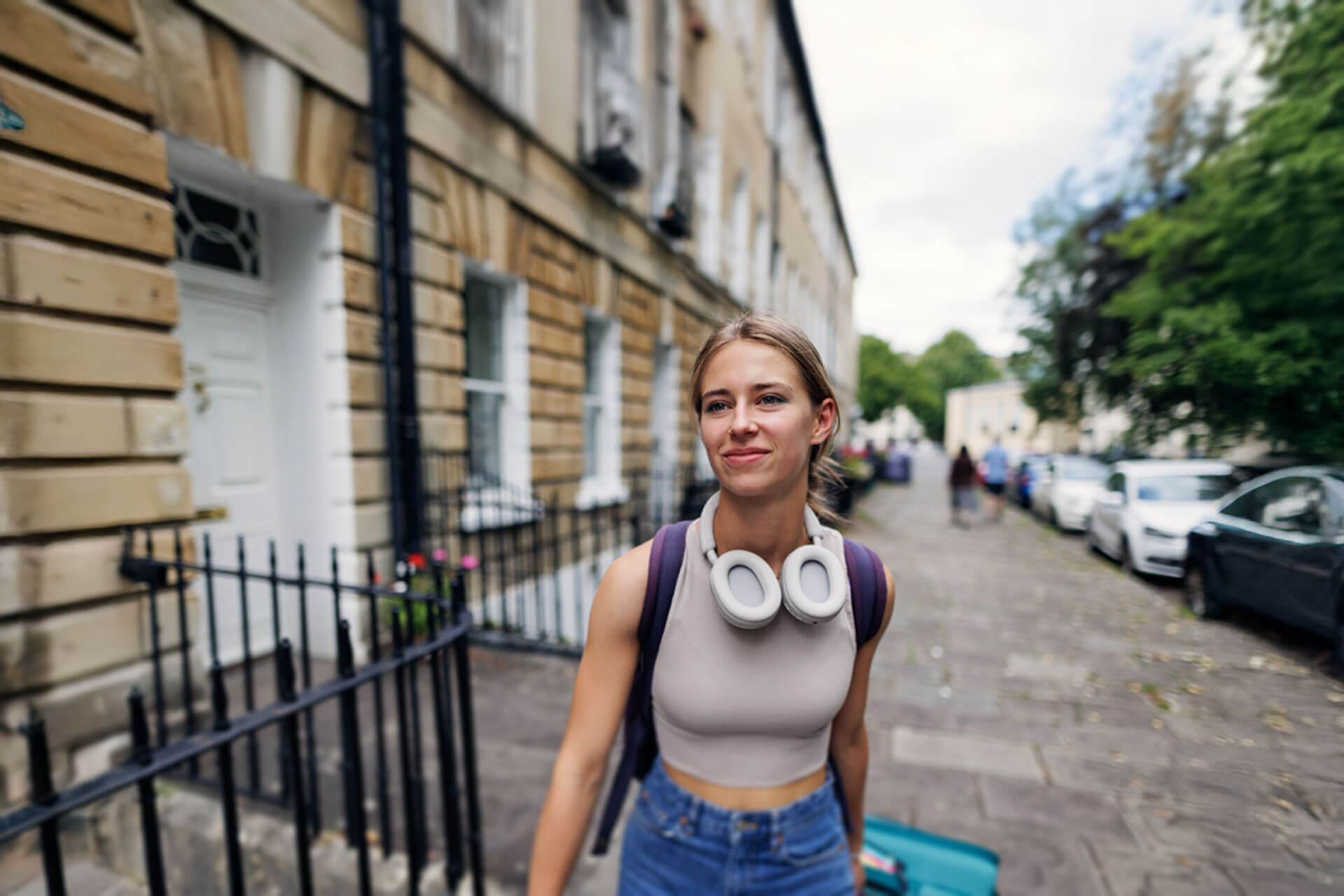Junge Frau mit Kopfhörern um den Hals und Rucksack läuft lächelnd durch eine typische englische Straße in Cambridge auf dem Weg zur Sprachschule.