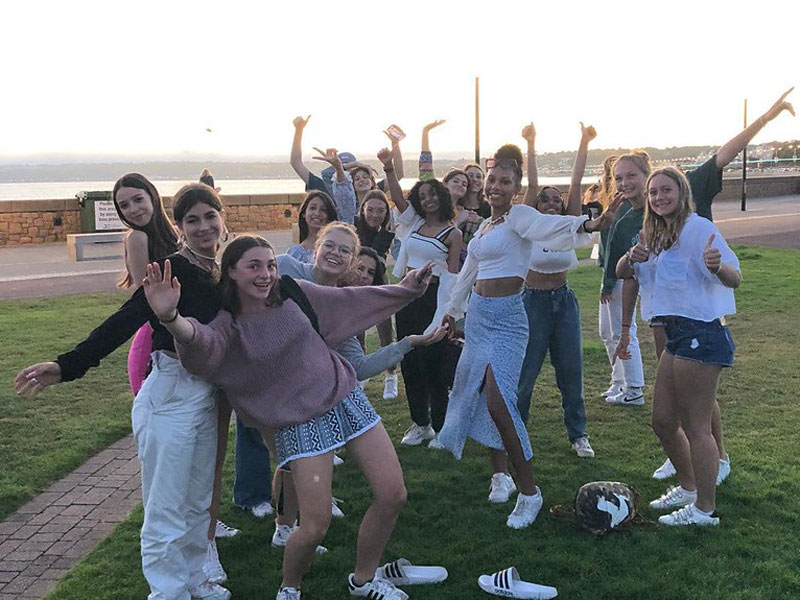 Gruppe junger Sprachschülerinnen feiert fröhlich am Meer bei Sonnenuntergang auf Jersey