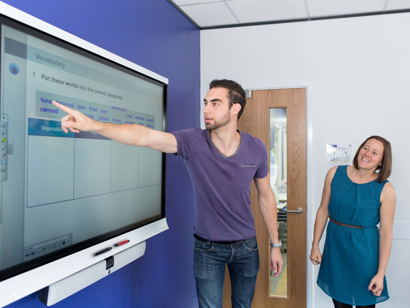 Lehrkraft erklärt interaktiv am Smartboard, während ein Teilnehmer Aufgaben an der Tafel löst