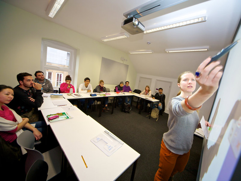 Lehrkraft erklärt Unterrichtsinhalte an der Tafel, während eine Gruppe von Sprachschüler:innen aufmerksam folgt