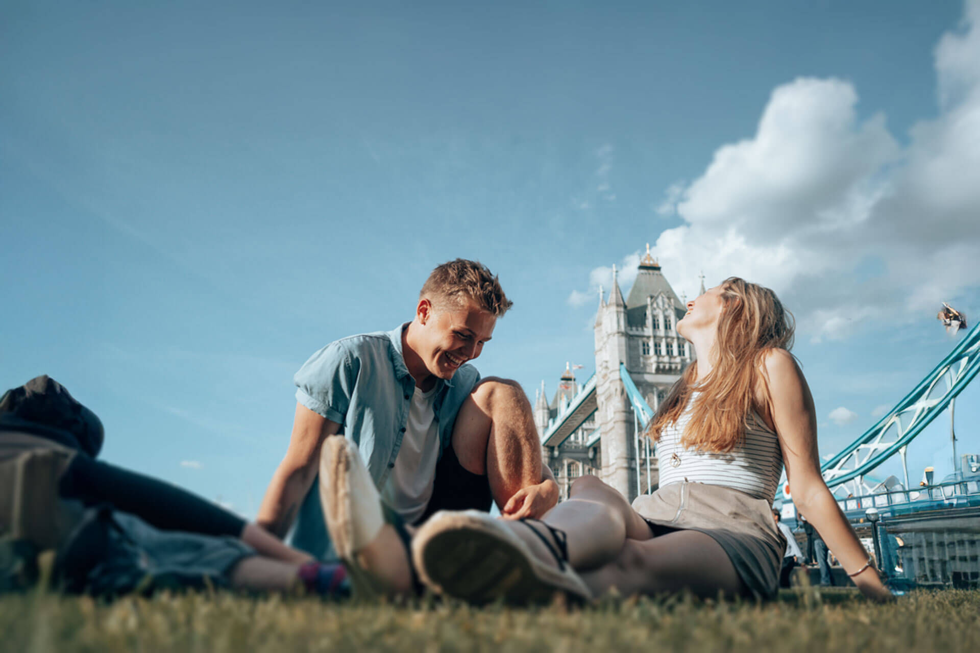Zwei Jugendliche sitzen entspannt auf der Wiese vor der Tower Bridge in London und genießen den Sonnenschein