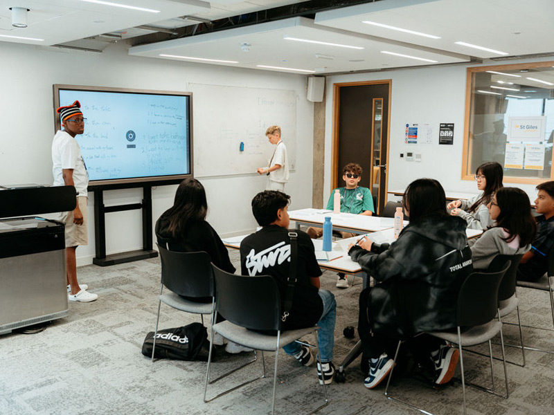 Lehrkraft erklärt Unterrichtsinhalte an Whiteboard und Smartboard vor einer Gruppe Jugendlicher