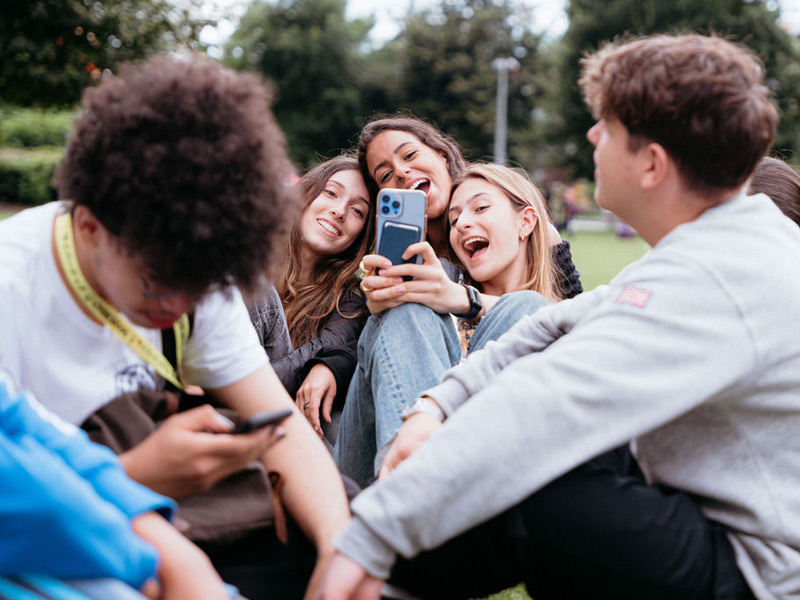 Gruppe junger Sprachschüler:innen sitzt im Park, lacht und macht gemeinsam Selfies