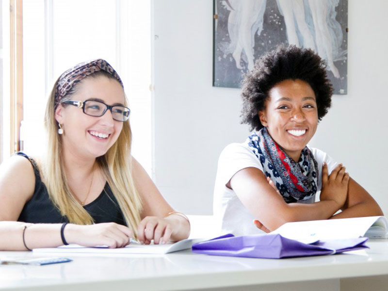 Zwei Sprachschülerinnen lachen während des Unterrichts in einem hellen Klassenraum