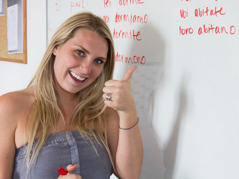 Teilnehmerin steht vor der Tafel mit italienischen Verben und zeigt Daumen hoch