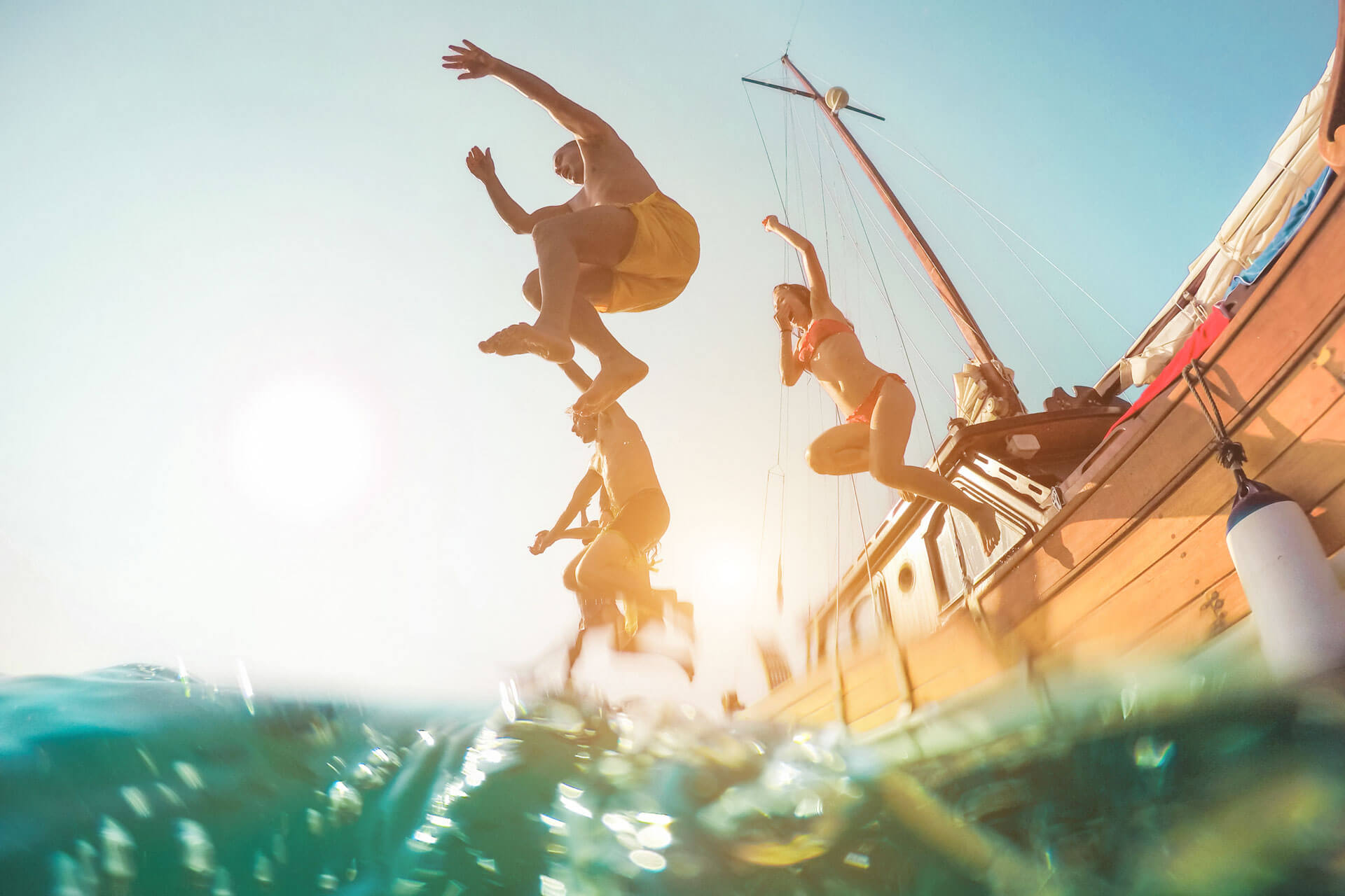 Gruppe junger Menschen springt fröhlich von einem Segelboot ins Meer an einem sonnigen Tag