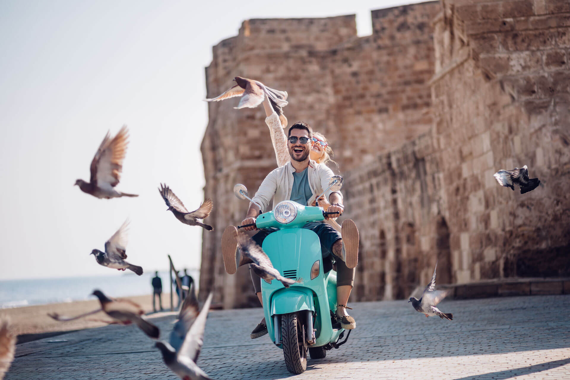Paar fährt lachend auf einer türkisfarbenen Vespa entlang der alten Stadtmauer von Sanremo, während Tauben auffliegen