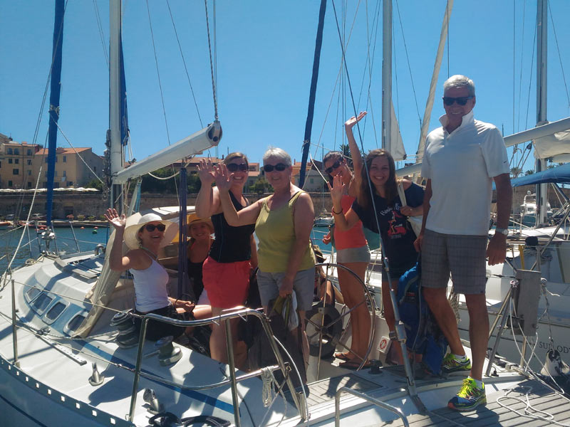 Teilnehmerinnen und Teilnehmer eines Ausflugs posieren fröhlich auf einem Segelboot im Hafen von Alghero