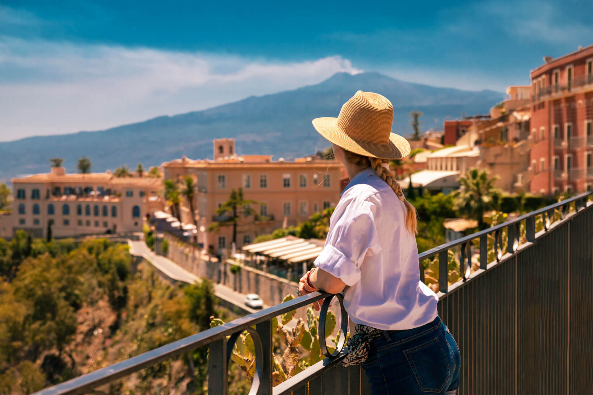 Junge Frau mit Strohhut blickt von einer Terrasse auf Taormina und den Ätna im Hintergrund