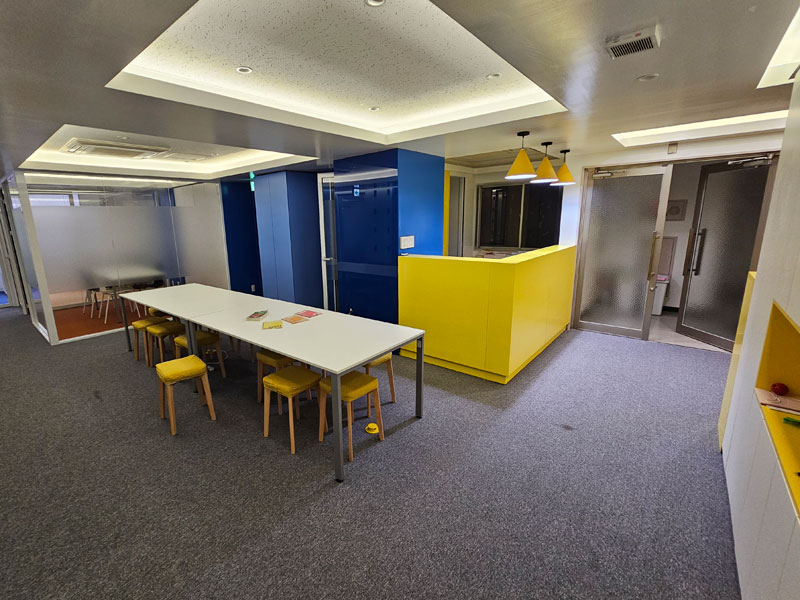 Moderner Aufenthaltsbereich der LTL Tokio Sprachschule mit gelber Rezeption und Gemeinschaftstischen