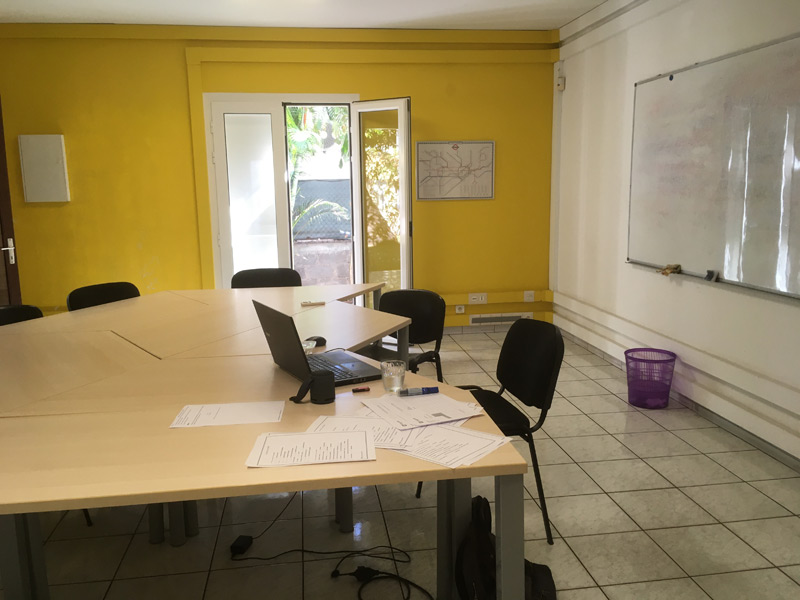 Heller Klassenraum mit großem Tisch, gelber Wand und geöffneter Tür zum Garten