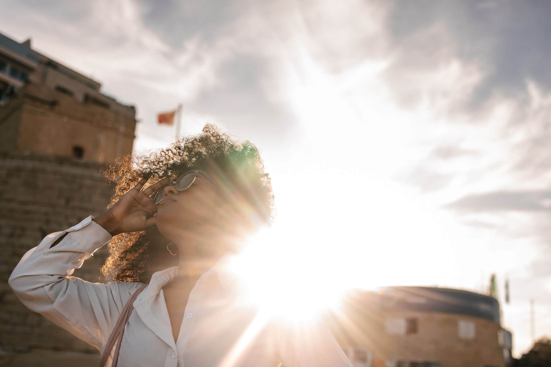 Junge Frau mit Sonnenbrille steht im Gegenlicht der Sonne vor historischen Gebäuden in Sliema, Malta