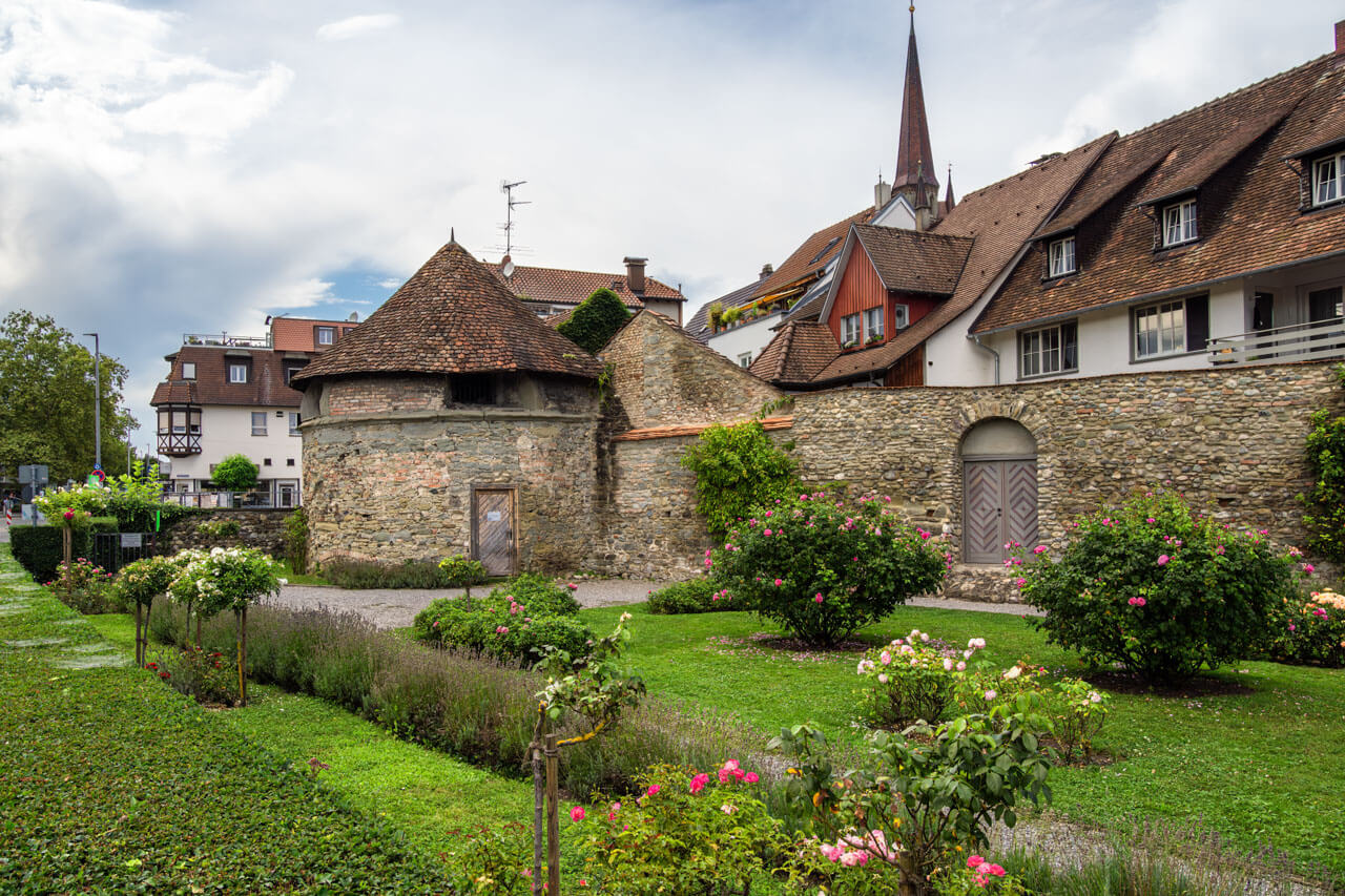 Historischer Stadtgarten mit Steinmauer, Rundturm und blühenden Rosenbeeten.