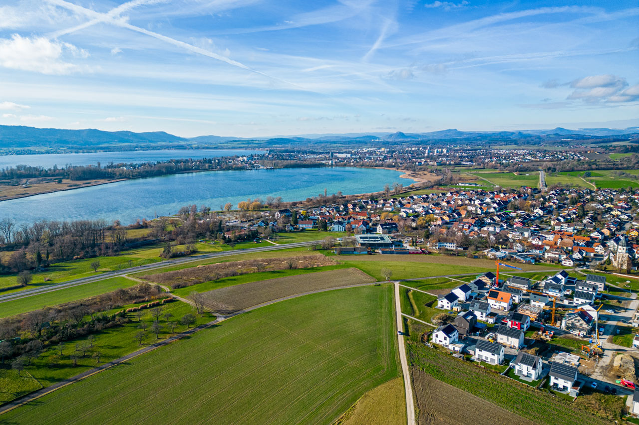 Luftaufnahme von Radolfzell mit Feldern, Wohnsiedlungen und Bodenseebuchten unter blauem Himmel.