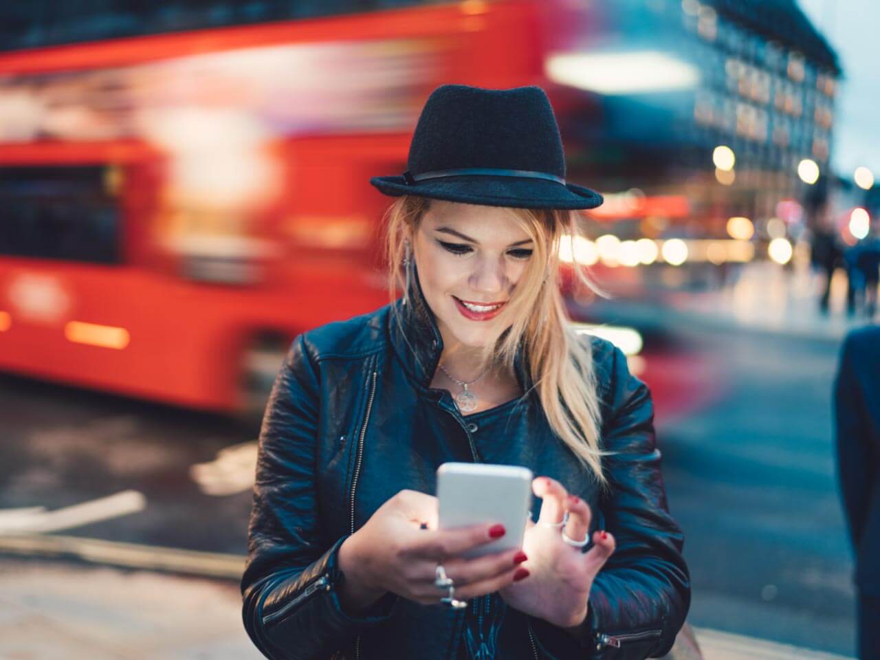 Junge Frau nutzt ihr Smartphone vor vorbeirauschendem roten Doppeldeckerbus in London Vokabeln und Alltagsausdrücke unterwegs während der Sprachreise üben