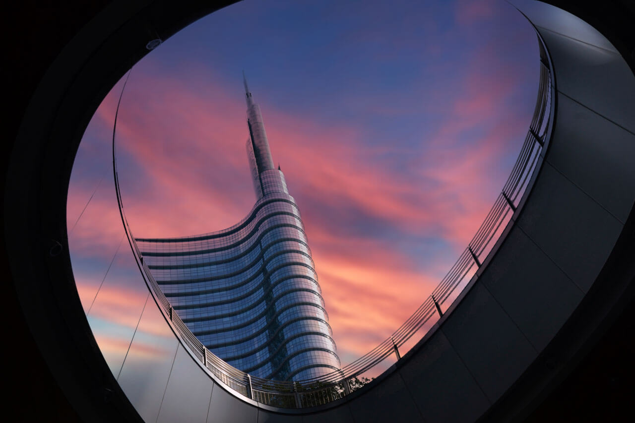 Moderner Wolkenkratzer in Mailand bei Sonnenuntergang, eingerahmt durch eine runde Architekturöffnung, Sinnbild für Konversation und Business Italienisch