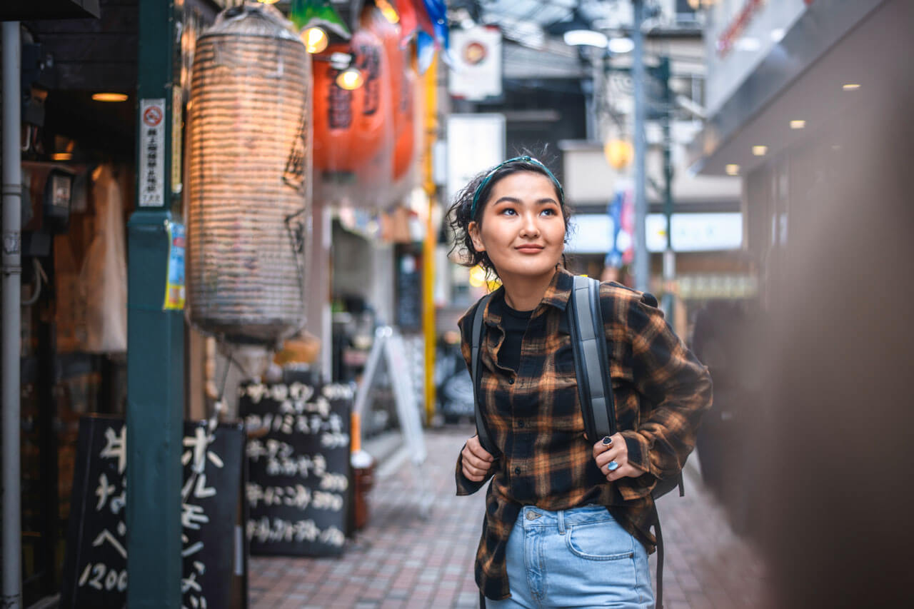 Junge Reisende mit Rucksack schlendert durch eine enge Gasse mit Laternen und Schildern