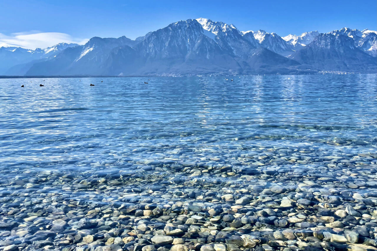Klares Uferwasser des Genfersees vor Alpen, Hörverstehen trainieren beim Spaziergang nach dem Sprachkurs