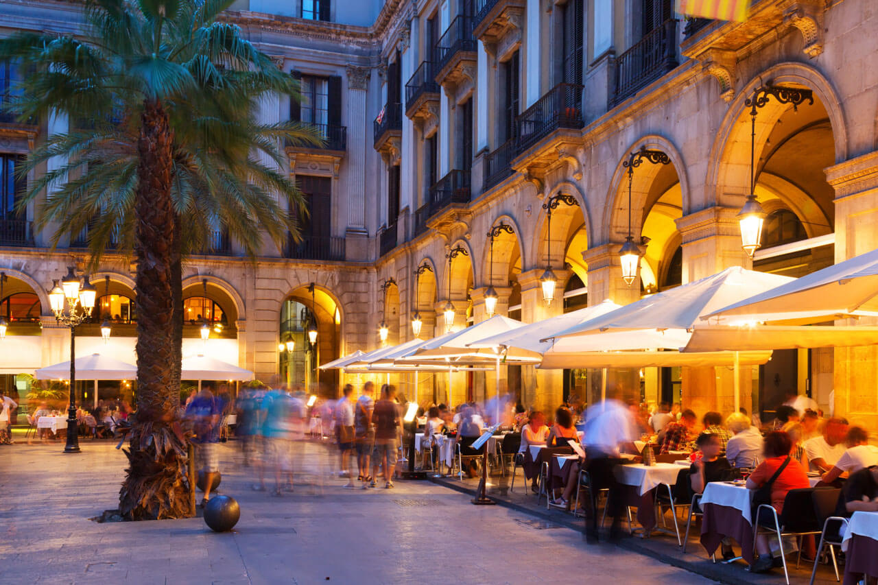 Plaça Reial am Abend mit Arkaden, Palmen und belebten Terrassenrestaurants.