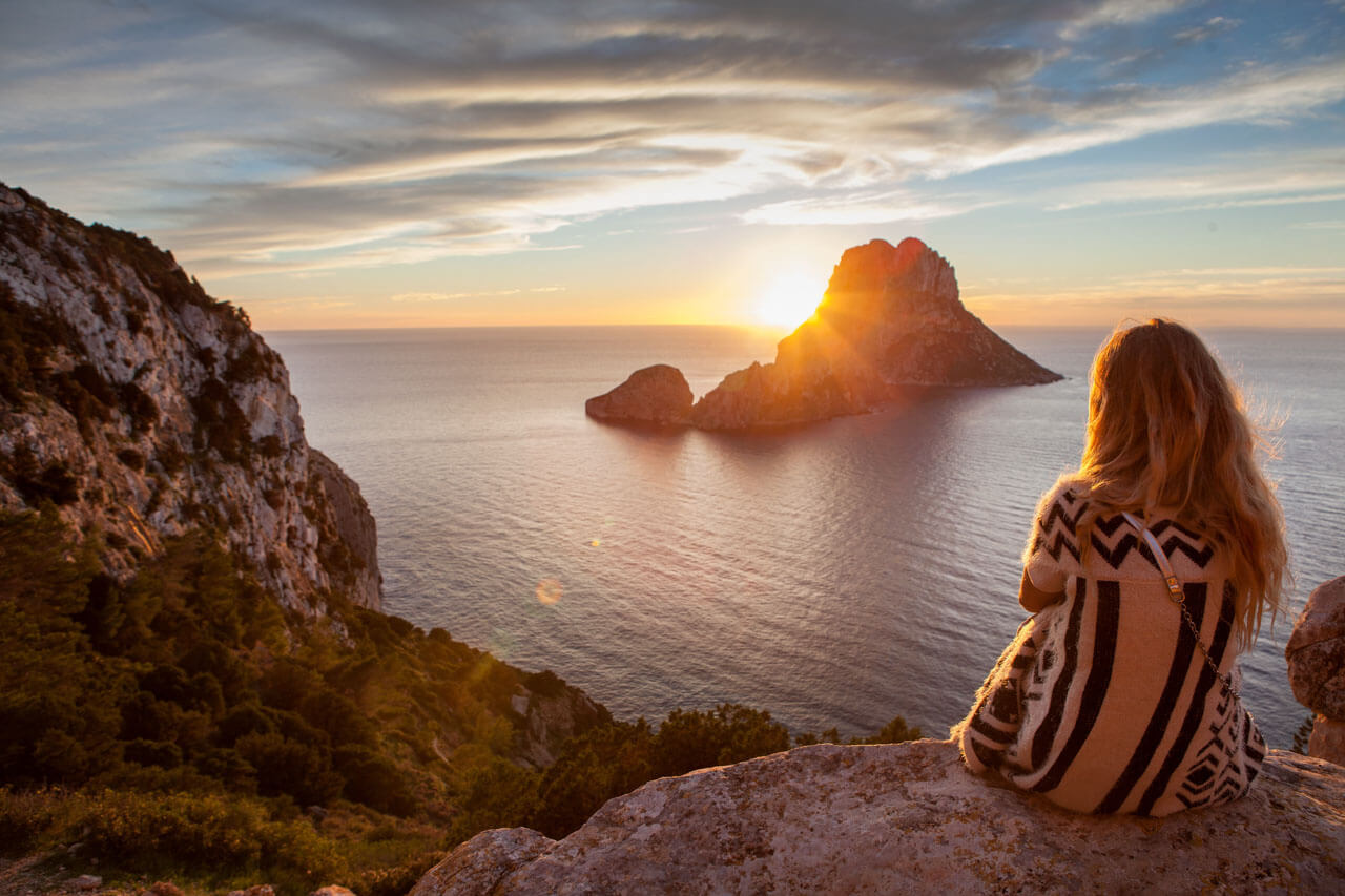 Sonnenuntergang mit Blick auf Es Vedrà – Person sitzt auf Felsen, dramatischer Himmel über dem glitzernden Mittelmeer.