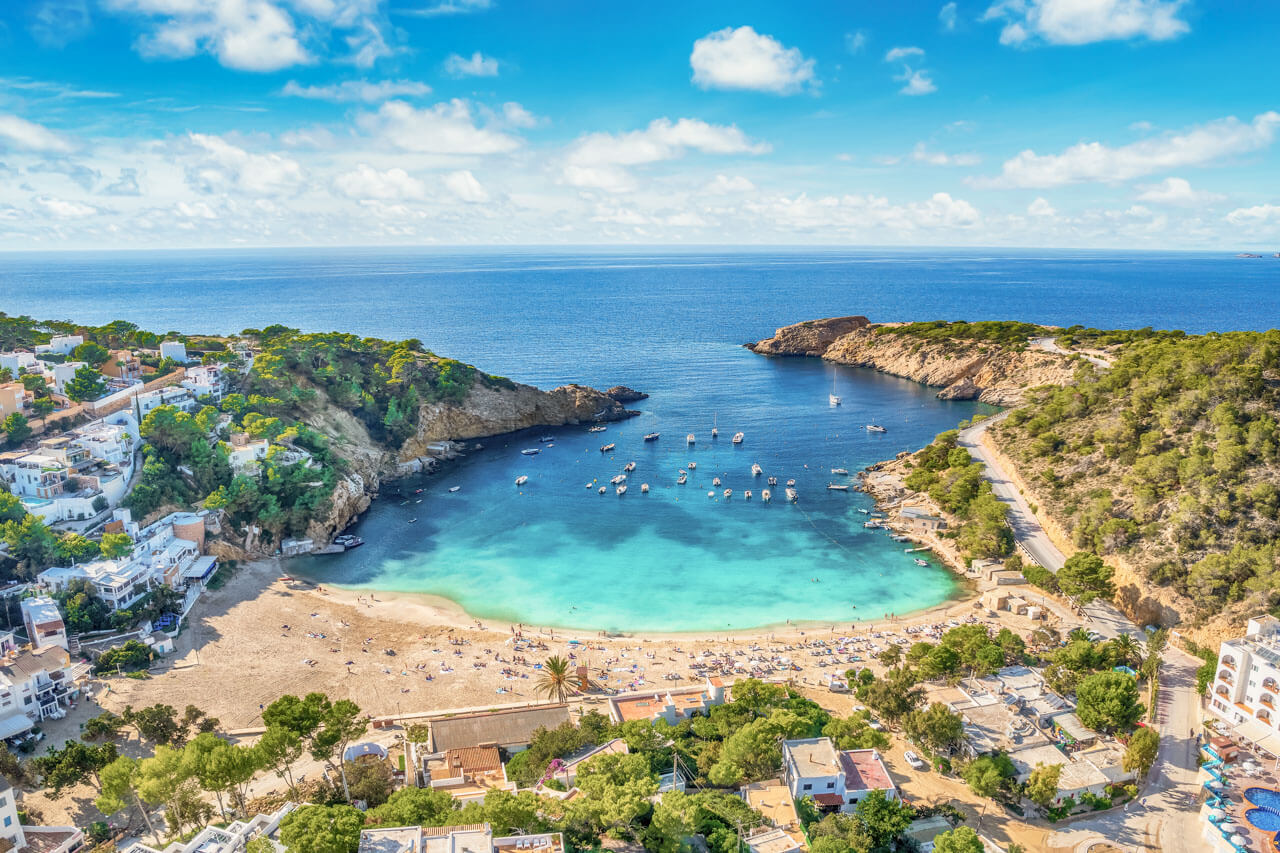 Luftaufnahme der Bucht Cala Saladeta/Cala Salada – türkisblaues Wasser, Boote, goldener Sandstrand und Pinienhänge.