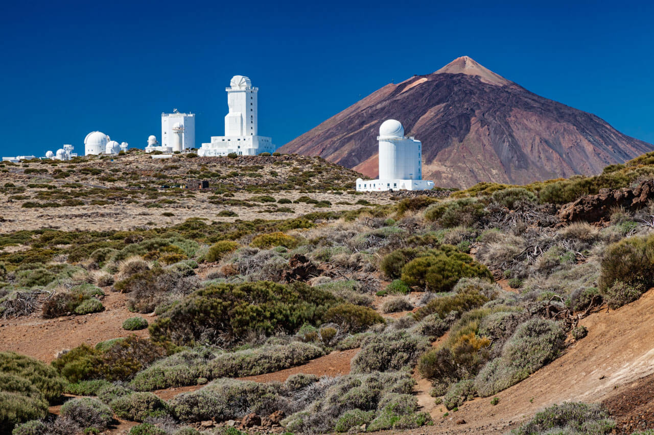 Teide-Observatorium mit weißen Teleskopen vor karger Vulkanlandschaft, Pico del Teide im Hintergrund unter tiefblauem Himmel.