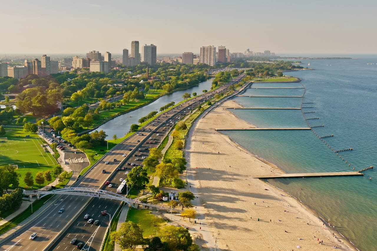 Luftaufnahme Sandstrand am Seeufer, Verkehrsweg daneben Sprachkurs mit Blick aufs Wasser.