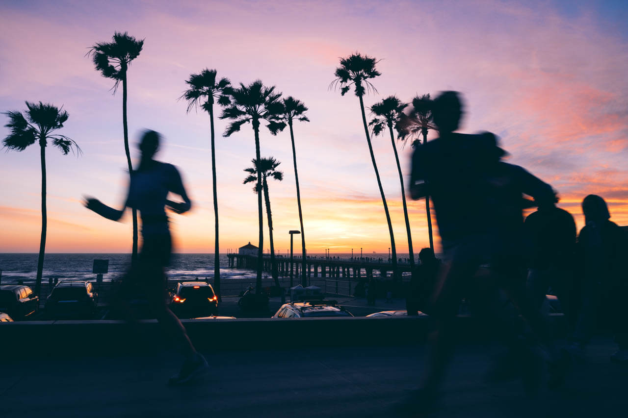 Jogger als Silhouetten vor Palmen und einem Pier bei farbigem Sonnenuntergang.