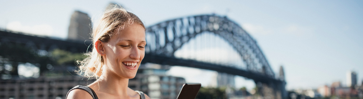 Junge Frau schaut lächelnd auf ihr Handy vor einer bekannten Brücke im Ausland.